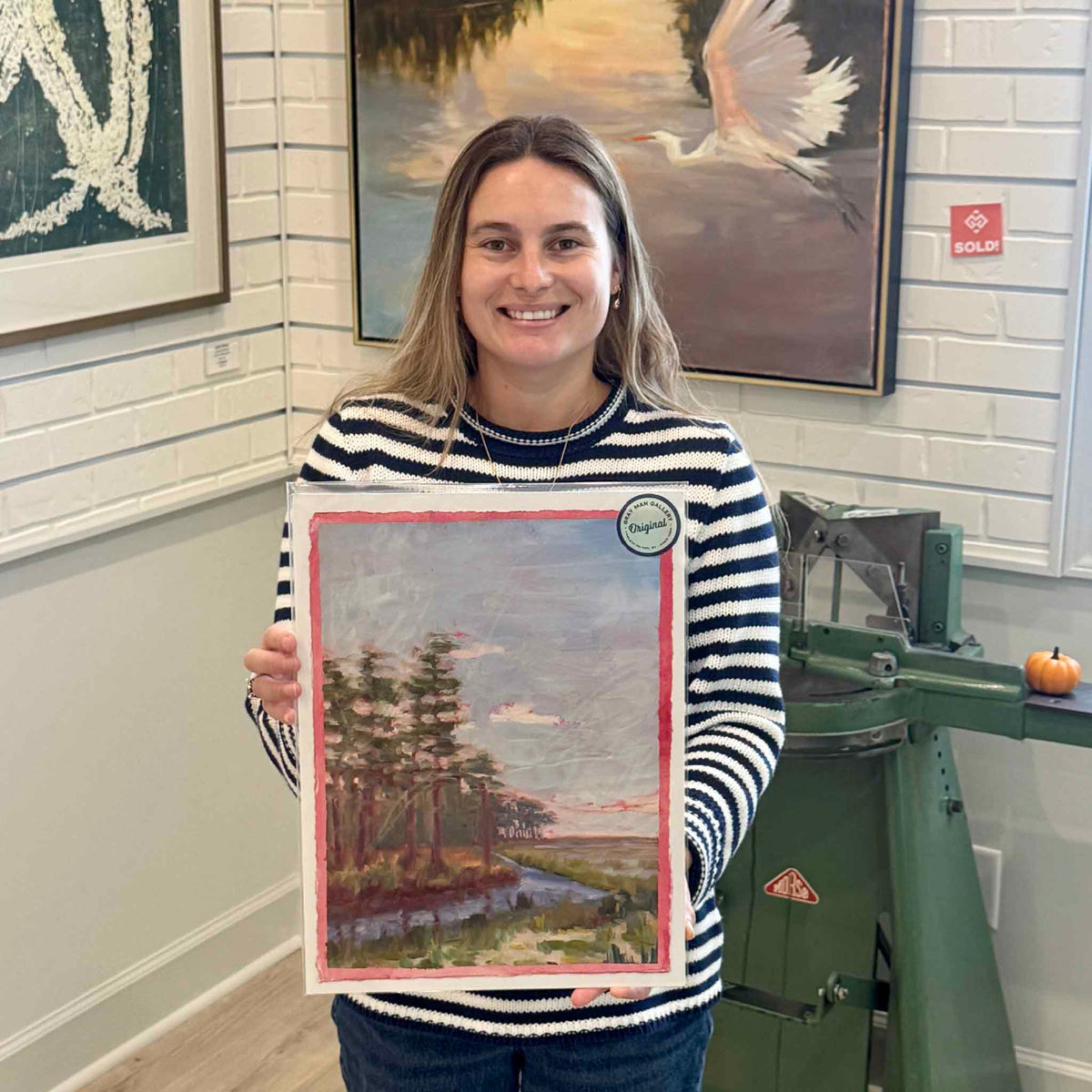 Gray Man Gallery team member holding South Island Pines #217, an original oil on paper by local artist Retta Dotterer, showing the scale of the pine and marsh landscape and its hand-painted pink border.