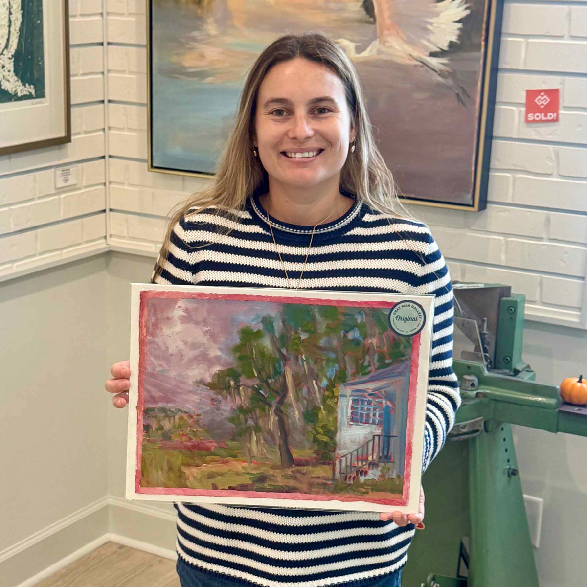 Gray Man Gallery team member holding Stormy Jamie’s #215, an original oil on paper by local artist Retta Dotterer, showing the scale, stormy Lowcountry scene and signature pink painted border.