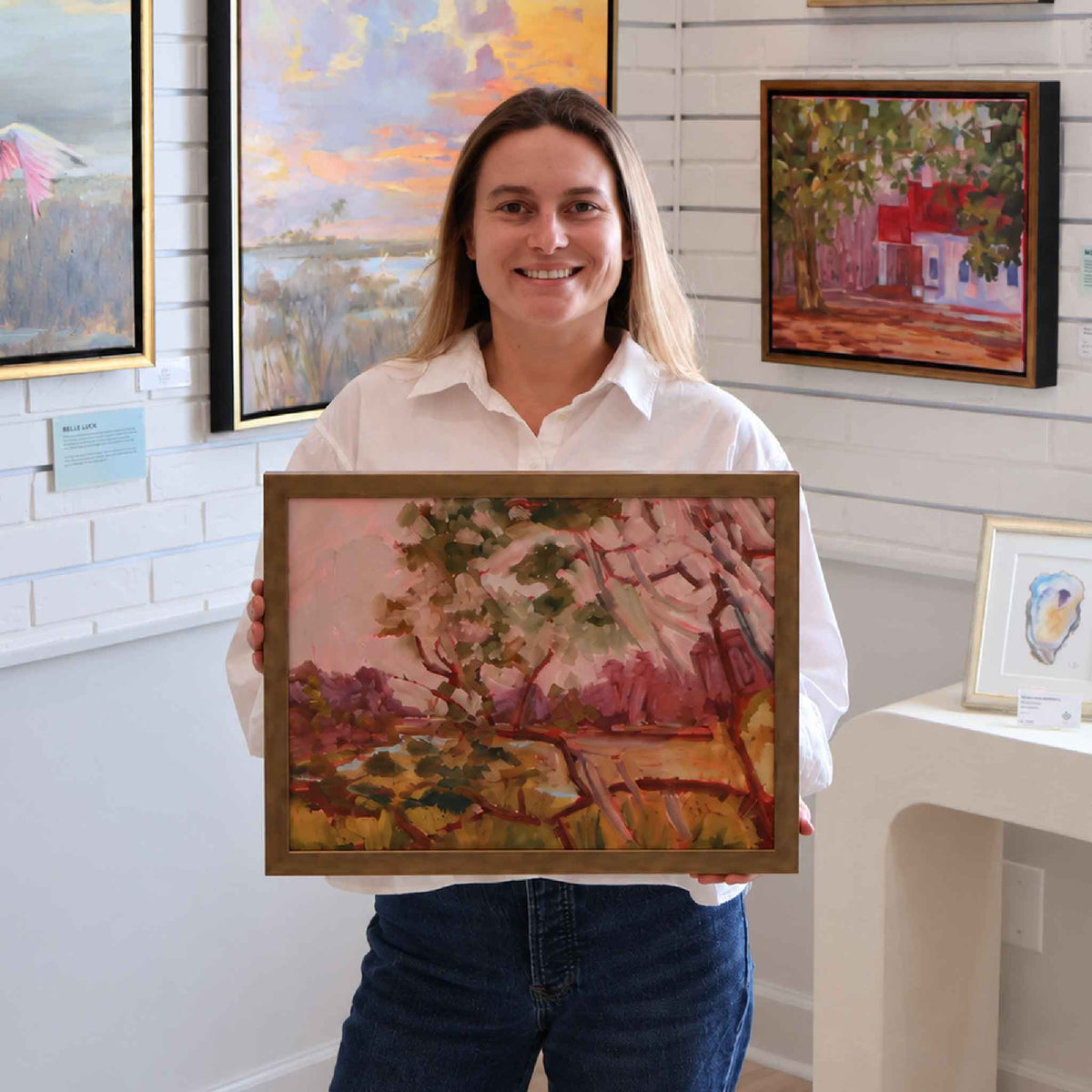 Gray Man Gallery team member holding Warm Afternoon #201, an original oil painting by local artist Retta Dotterer featuring colorful Lowcountry live oaks in expressive pink, lavender and green brushwork, shown for scale at the Pawleys Island gallery.
