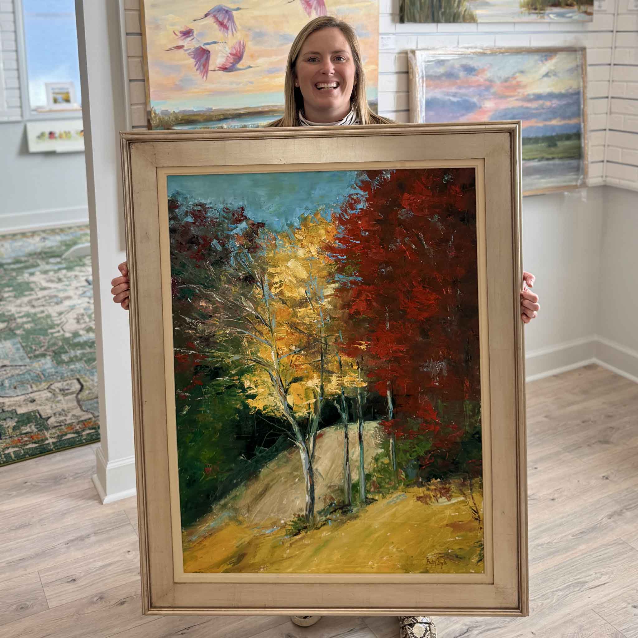 Local artist Betsy Boyle’s original oil painting “Rich Hues” held by Gray Man Gallery team member Bordy inside the Pawleys Island gallery, showcasing professionally framed Lowcountry landscape artwork with bold autumn colors, expressive brushwork, and Southern coastal influence