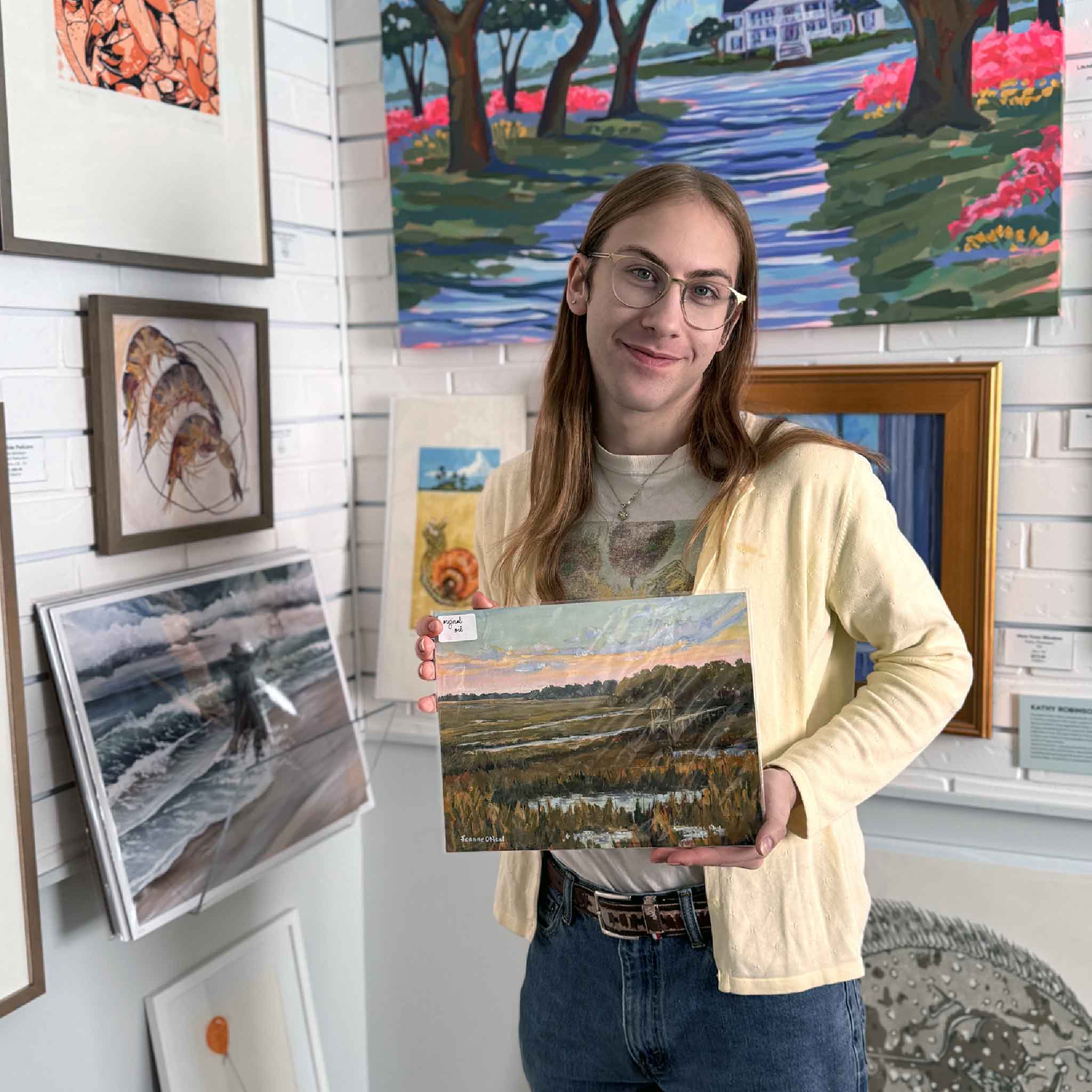 A gallery staff member holds Jeanne O’Neal’s Salt + Light #2 oil painting, showing its size and the warm dusk colors of the Lowcountry marsh scene.