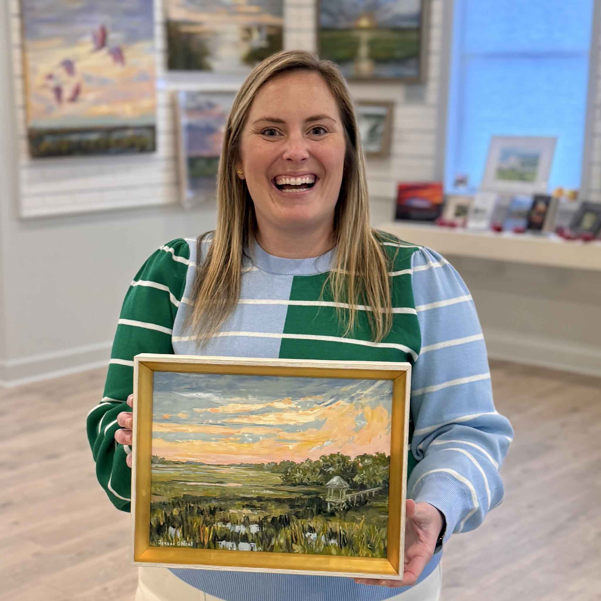 Gray Man Gallery team member holding “Salt + Light #3” original oil painting by Jeanne O’Neal, framed Lowcountry marsh sunset artwork inside the Pawleys Island gallery