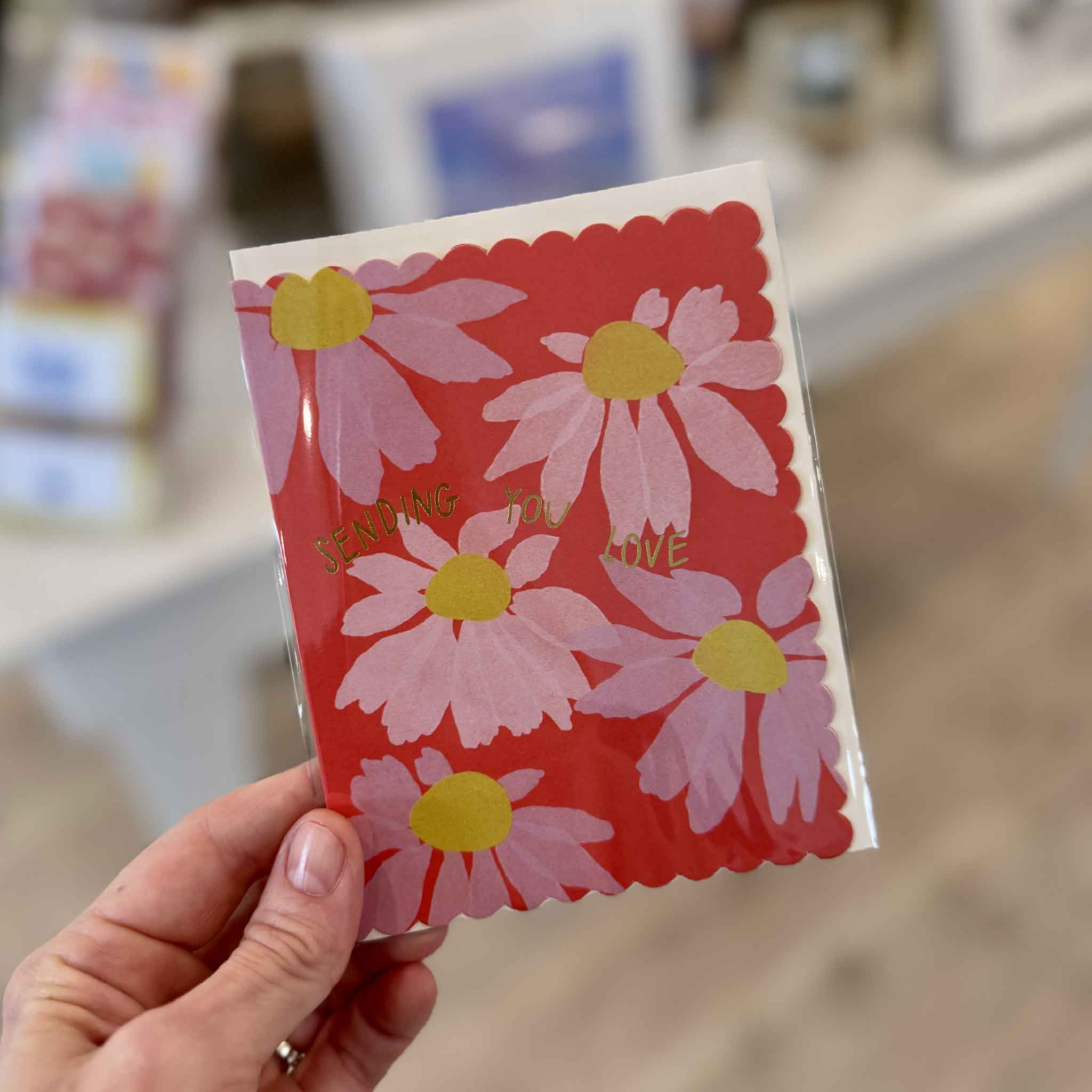 Hand holding the Sending You Love floral greeting card with pink coneflowers, sunny yellow centers, coral-red background, metallic gold foil message and decorative scalloped edges, a colorful thinking-of-you and just-because card available at Gray Man Gallery in Pawleys Island, South Carolina.