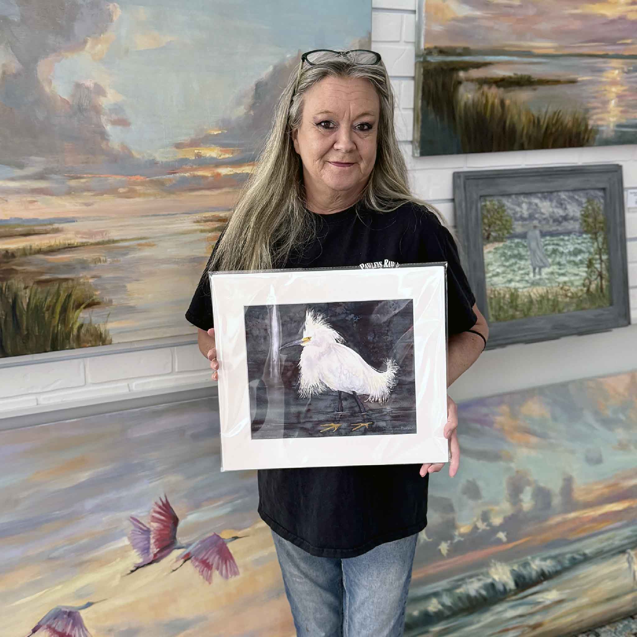 A Gray Man Gallery team member holds Shore Bird 3 by Mimi Beaver, an original watercolor of a white shorebird standing in dark tidal water, donated to benefit Salty Dogs Senior Animal Rescue.