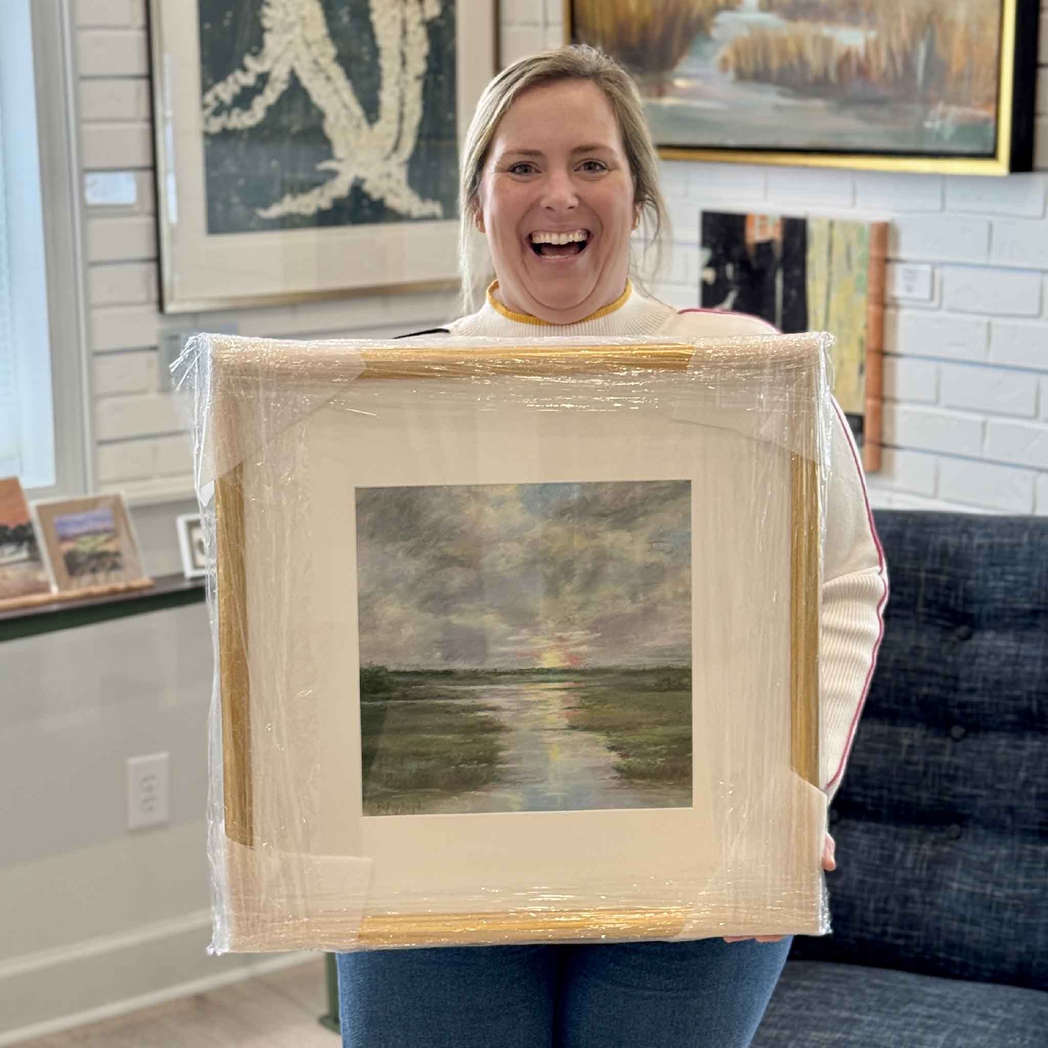 Gray Man Gallery team member holding “Soft Farewell,” an original pastel by local artist Betsy Boyle depicting a sunset over a South Carolina Lowcountry marsh, professionally framed and displayed in Pawleys Island art gallery