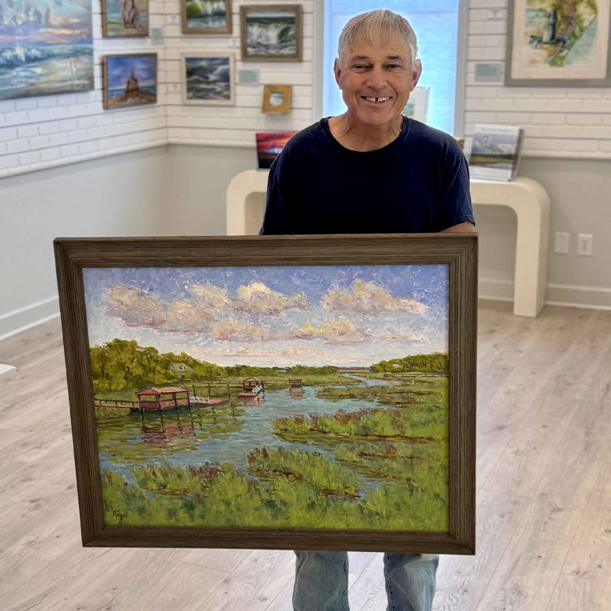 Local artist Michael Rogan holding Spring Greens original oil painting at Gray Man Gallery, showing scale of Pawleys Island marsh scene with creek, docks, and vibrant green grasses