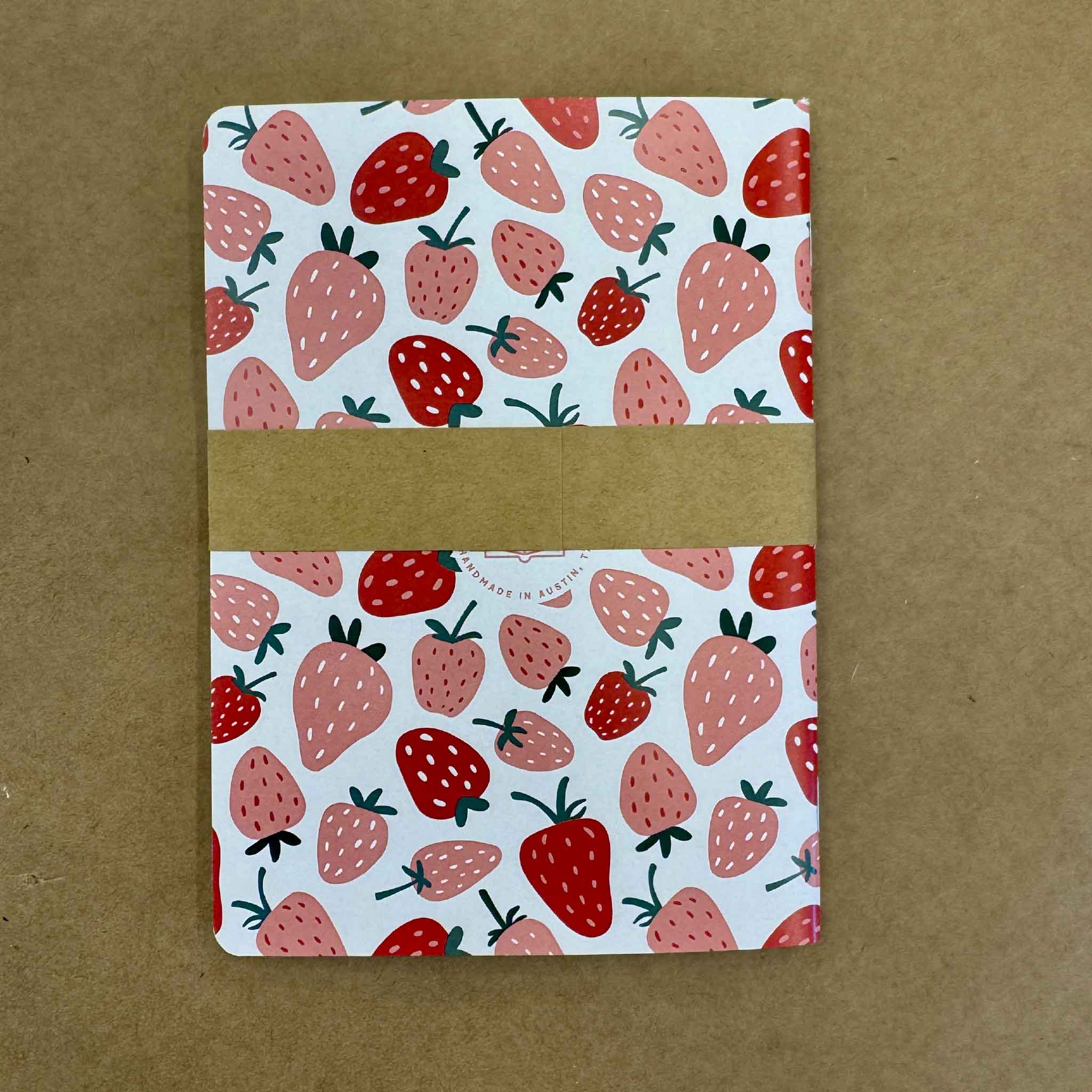 Strawberry Fields lined notebook shown with branded belly band packaging. The 5x7 journal features a colorful strawberry print cover, lined interior pages, and a durable cardstock cover, making it a fun gift for writers, students, teachers, and stationery lovers.