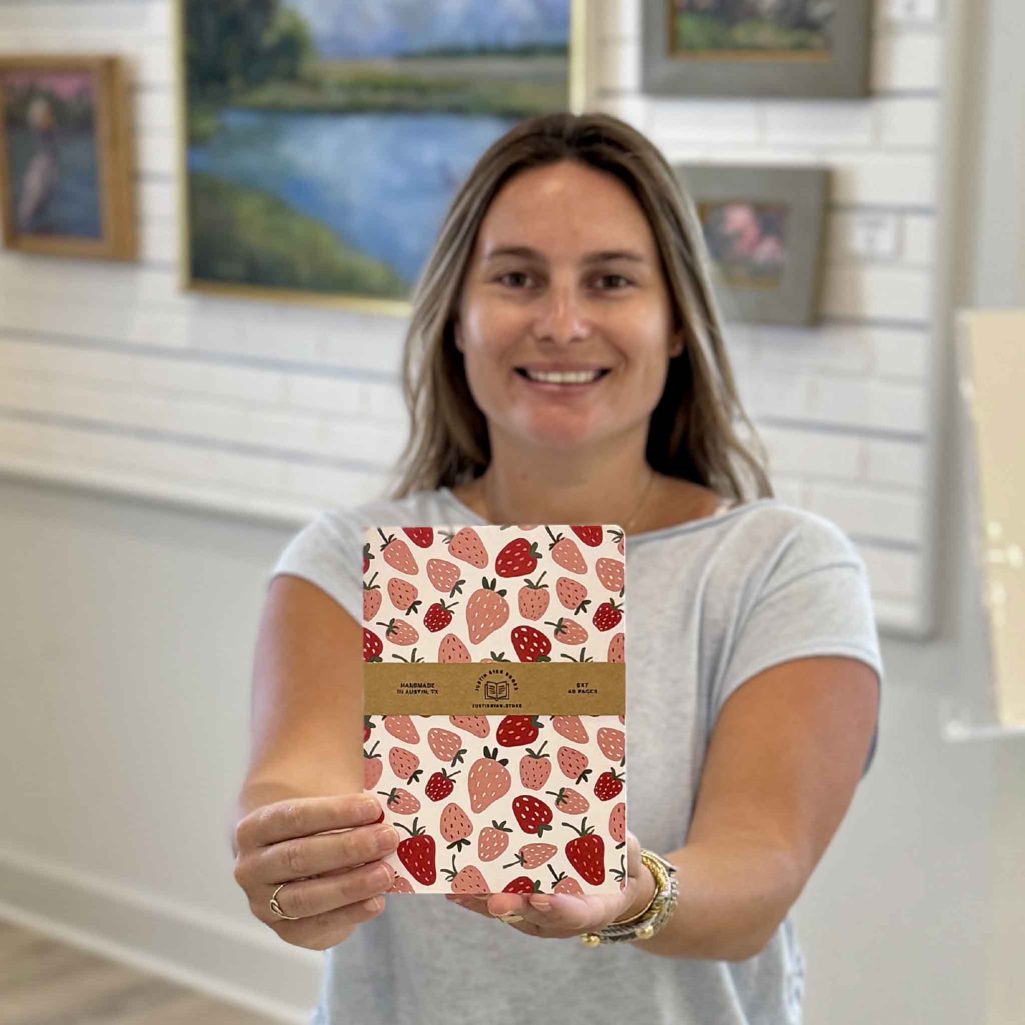Gray Man Gallery team member Kyra holding the Strawberry Fields 5x7 lined notebook in the gallery. This cheerful journal features an all-over strawberry pattern and is perfect for journaling, note-taking, planning, and everyday writing.