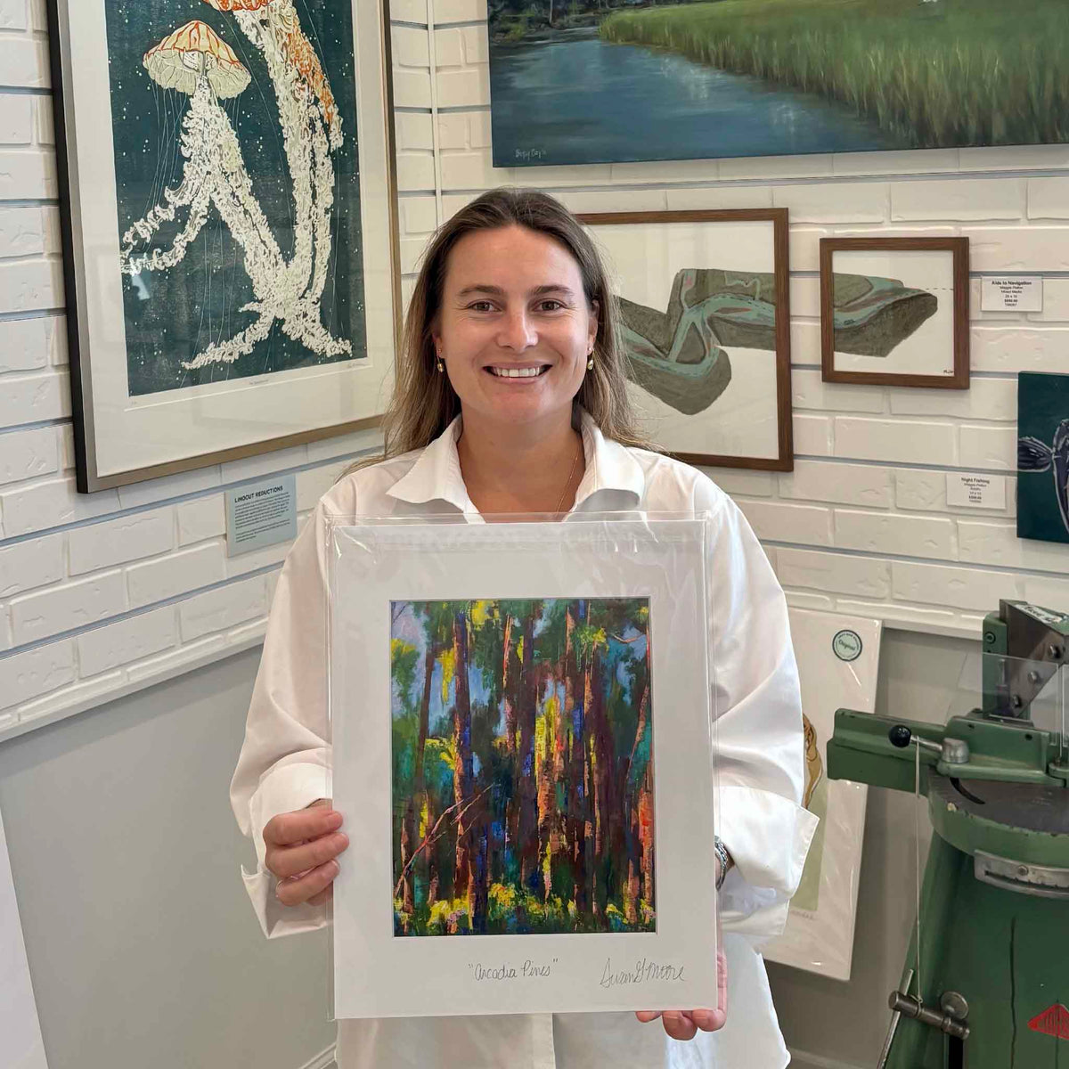 Kyra holding Susan Moore’s Arcadia Pines fine art giclée print inside Gray Man Gallery in Pawleys Island, South Carolina, featuring an expressive woodland landscape of tall pine trees in vibrant green, blue, gold and violet, professionally matted and personally titled and signed by Susan Moore.