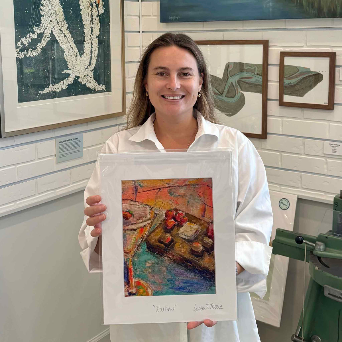 Kyra holding Gather by Susan Moore, a professionally matted fine art giclée print of a colorful dining table still life, hand-titled and signed by the artist and photographed inside Gray Man Gallery in Pawleys Island, South Carolina.
