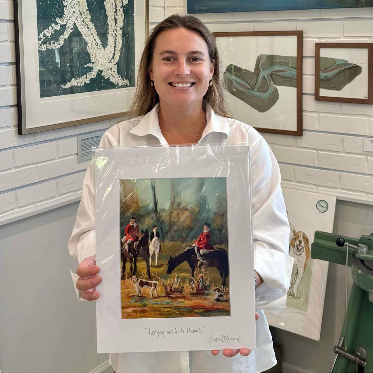 Kyra holding Susan Moore’s Hanging with the Hounds fine art giclée print inside Gray Man Gallery in Pawleys Island, South Carolina, featuring horseback riders in traditional hunt attire with hounds in a colorful equestrian scene, professionally matted and personally titled and signed by Susan Moore.