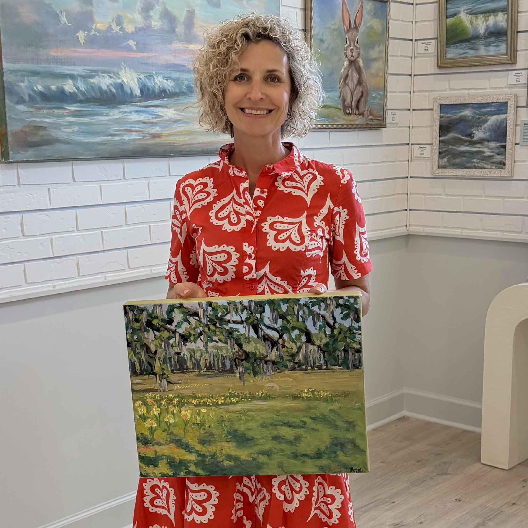 Susan Moore holding Spring Under the Oaks original acrylic landscape painting inside Gray Man Gallery Pawleys Island showing scale of gallery wrapped canvas with live oaks and blooming flowers