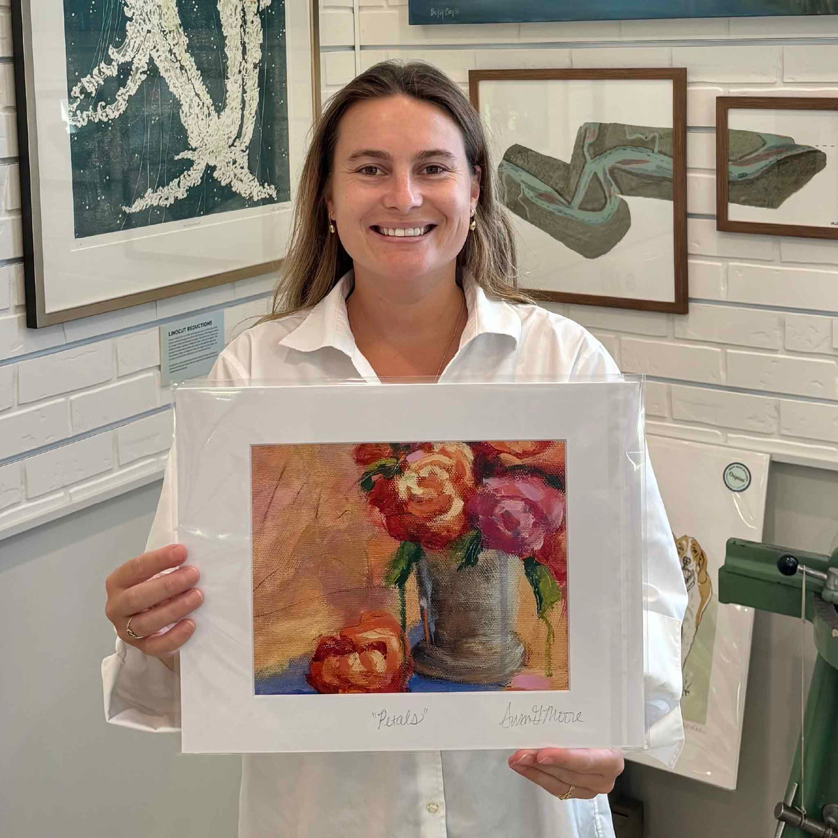 Kyra holding Susan Moore’s Petals floral giclée print inside Gray Man Gallery in Pawleys Island, South Carolina, featuring a colorful still life of pink, coral and red roses in a vase, professionally matted and personally titled and signed by Susan Moore.