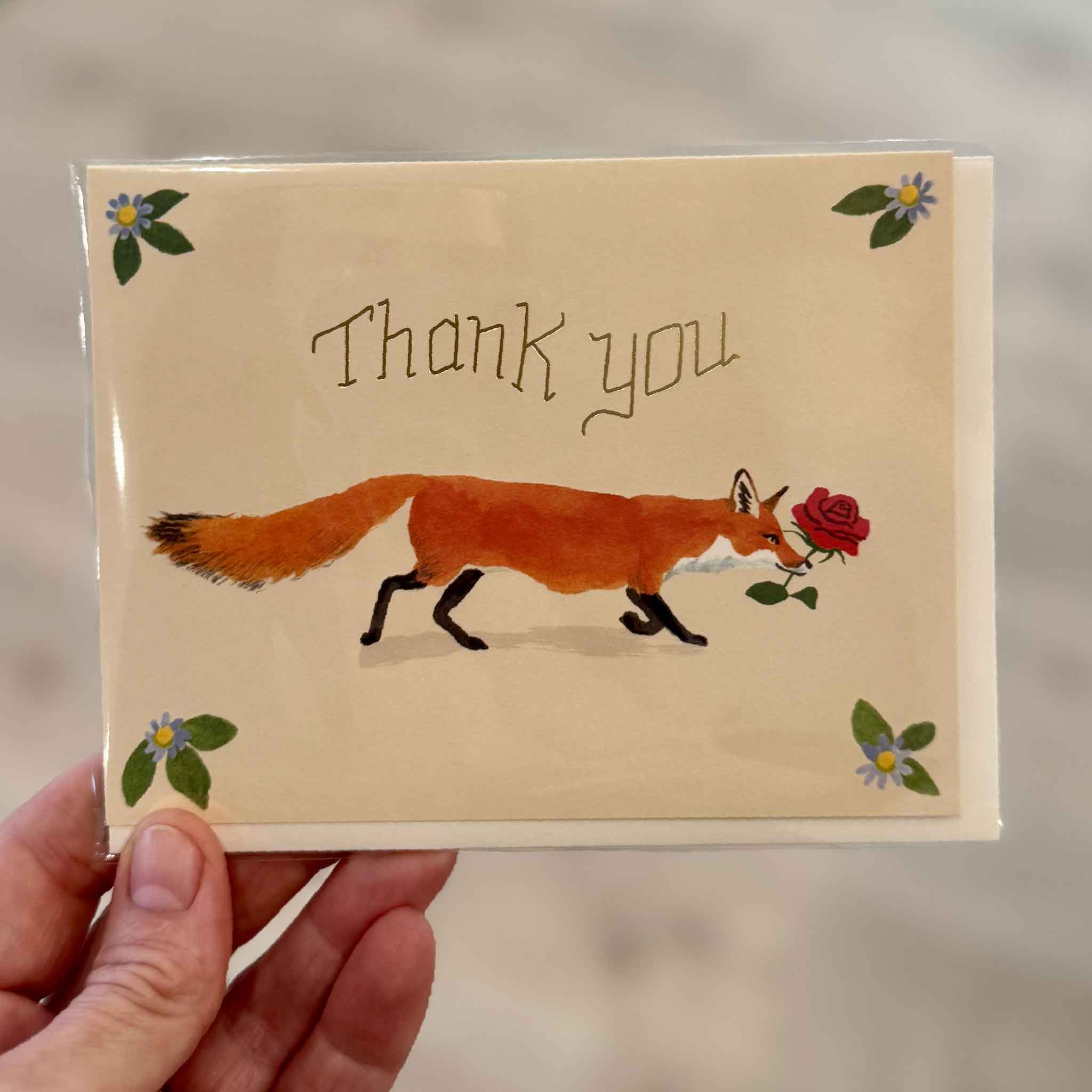 A hand holds the Thank You Fox greeting card by Danielle Kroll inside Gray Man Gallery in Pawleys Island, South Carolina. Featuring a charming red fox carrying a rose, delicate floral accents, and shimmering gold foil lettering, this blank-inside Red Cap Cards greeting card is an elegant way to thank friends, family, teachers, hosts, clients, or anyone deserving of a heartfelt note of appreciation.