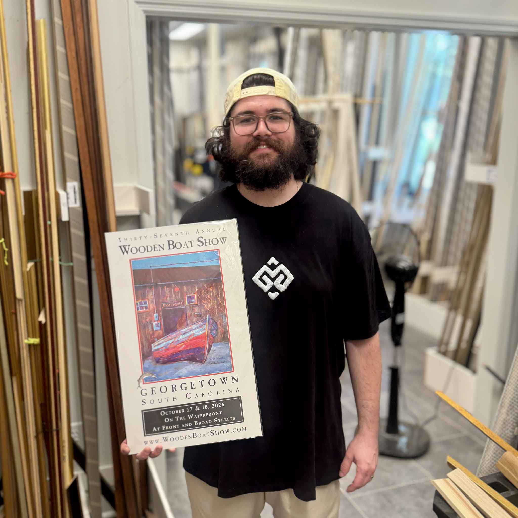 Peyton holds an unframed 2026 Georgetown Wooden Boat Show limited-edition artist-remarqued poster by Vida Miller inside the custom framing workshop at Gray Man Gallery in Pawleys Island, South Carolina; one of only ten posters individually embellished with an original hand-painted sturgeon, then signed and numbered by the artist.