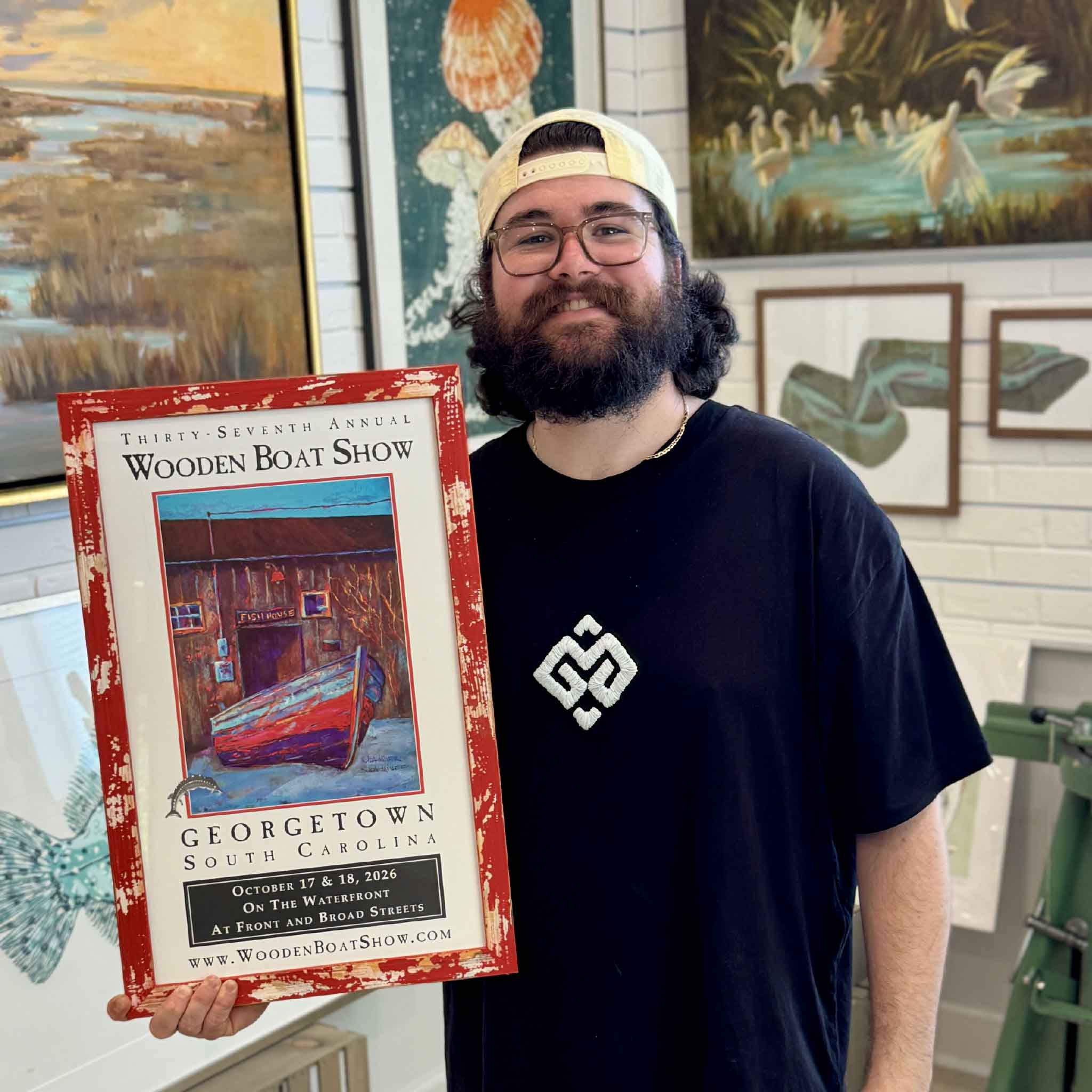 Peyton holds the framed 2026 Georgetown Wooden Boat Show limited-edition artist-remarqued poster by Vida Miller inside Gray Man Gallery in Pawleys Island, featuring Miller’s Cathou Fish House artwork and an original hand-painted sturgeon remarque; this custom red distressed frame was personally selected by Vida Miller for the poster, and is available for sale separately.