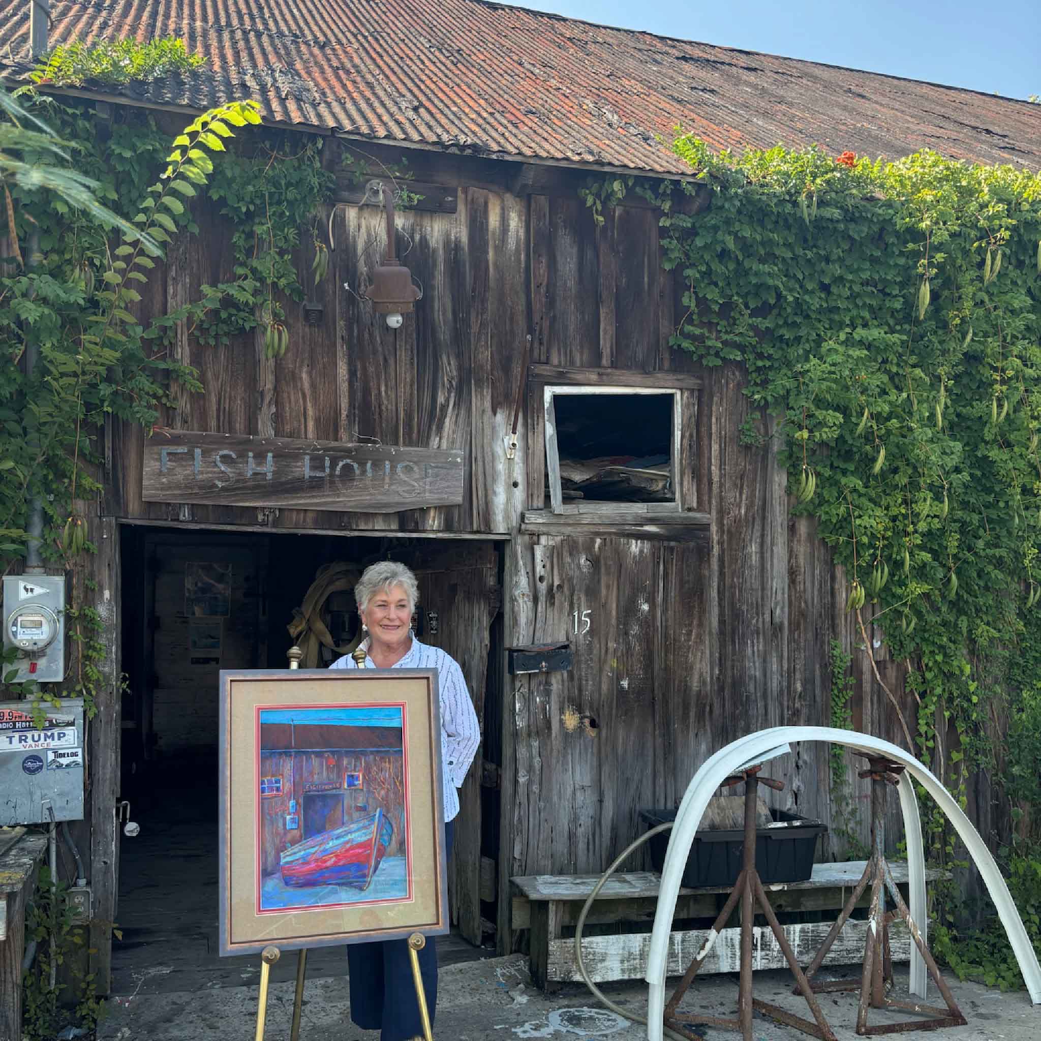 South Carolina artist and Gray Man Gallery founder Vida Miller stands outside the historic Cathou Fish House in Georgetown, SC, with her original artwork that became the 2026 Georgetown Wooden Boat Show poster, the same historic building whose carved sturgeon inspired her exclusive series of ten hand-painted artist-remarqued posters.