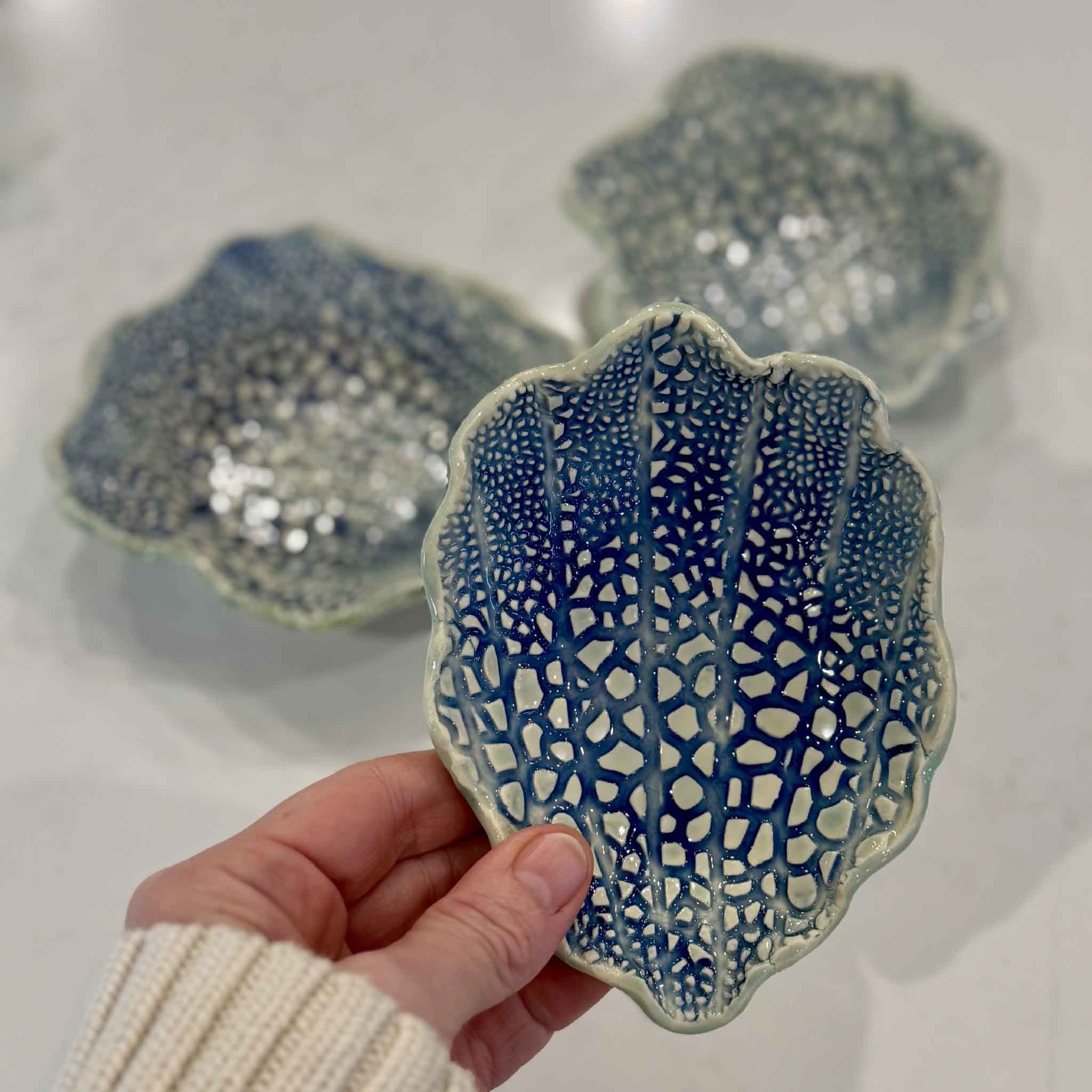 A hand holds Vida Miller's handmade Coral Shell Dish, showcasing the intricate coral-inspired texture, vibrant blue glaze, and handcrafted details of this one-of-a-kind coastal pottery piece available at Gray Man Gallery in Pawleys Island.