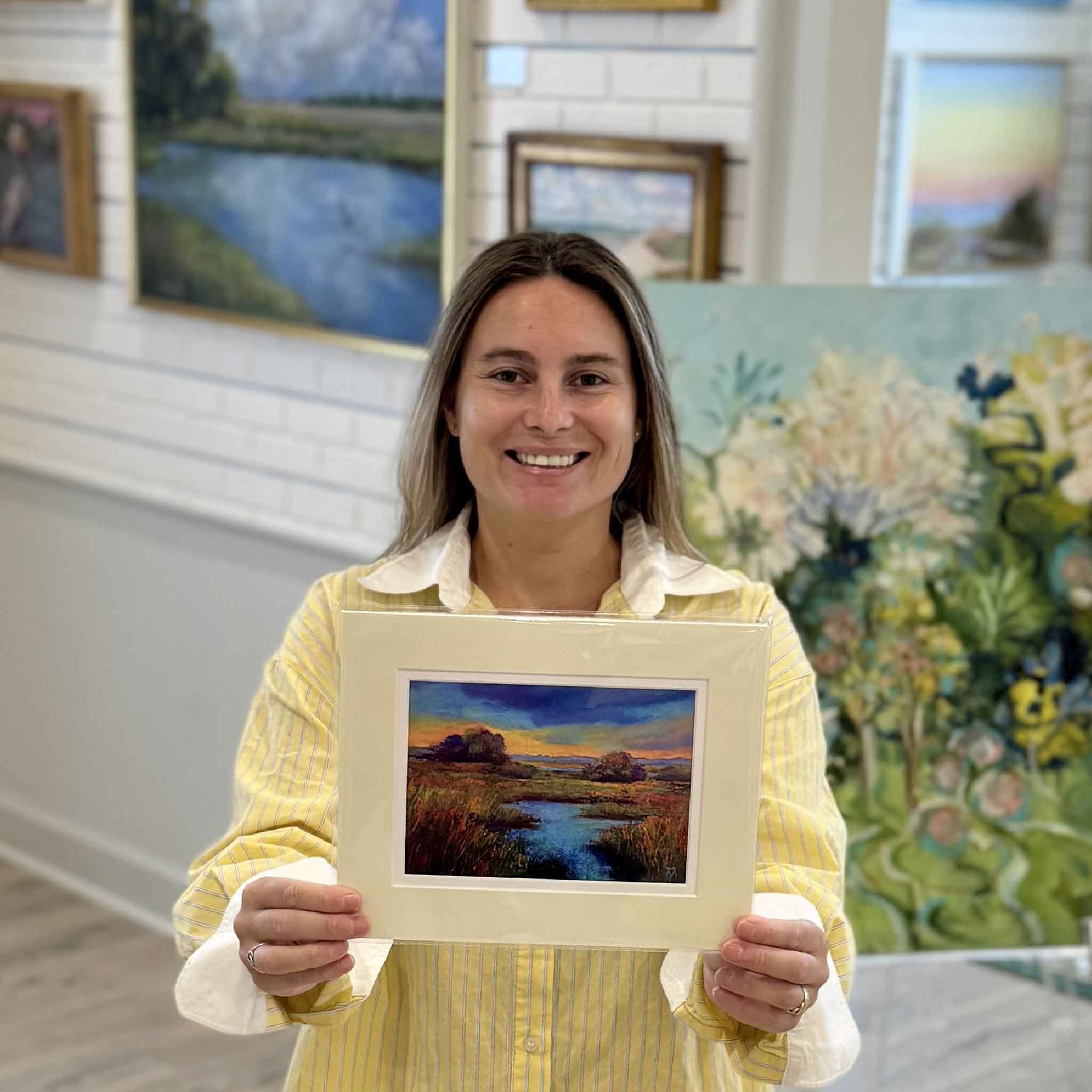 Kyra holds Marsh #6, a double-matted Lowcountry marsh print by Pawleys Island artist Vida Miller inside Gray Man Gallery. Reproduced from Miller's original pastel painting, this colorful 8×10 artwork features vibrant blue waterways, golden marsh grasses, and a dramatic coastal sky while fitting any standard ready-made frame.