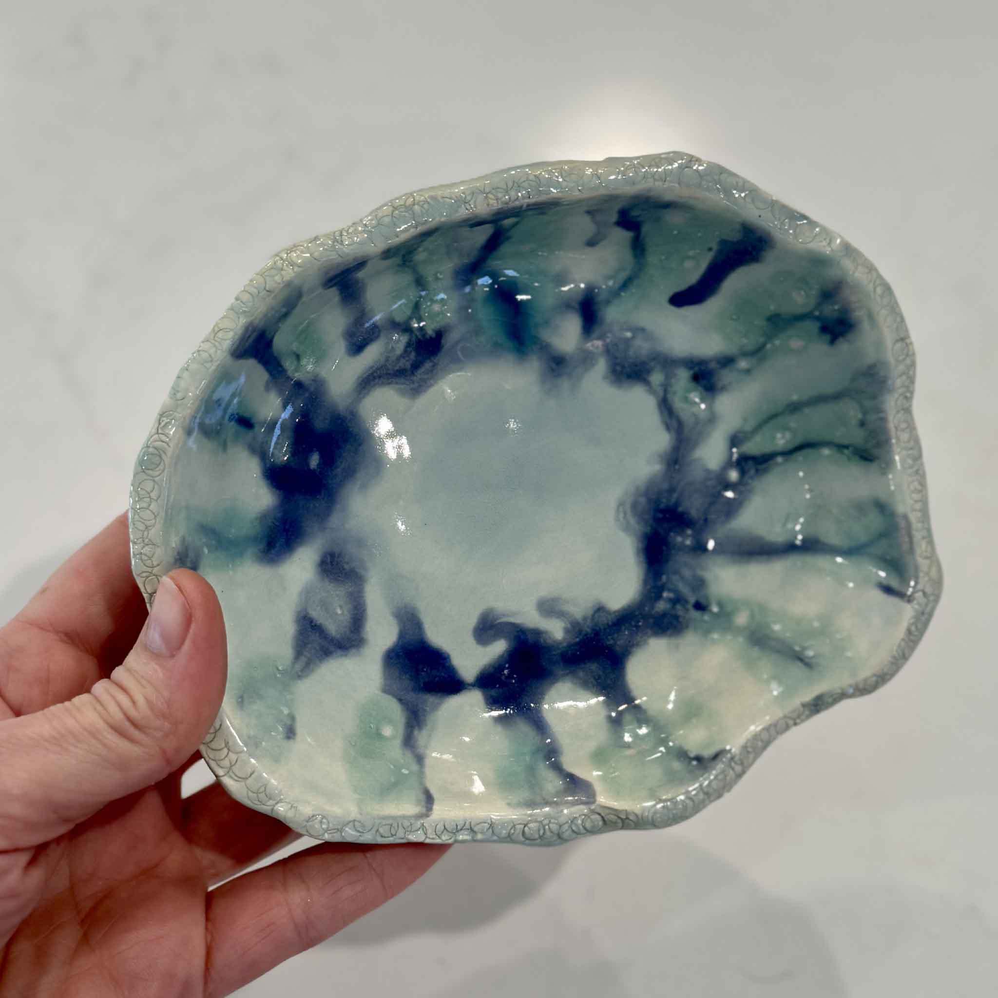 Hand holding the handmade Marsh Drift Bowl by Vida Miller for scale at Gray Man Gallery in Pawleys Island, South Carolina. This organic ceramic bowl features flowing blue-green and indigo glaze with a softly sculpted handmade edge.