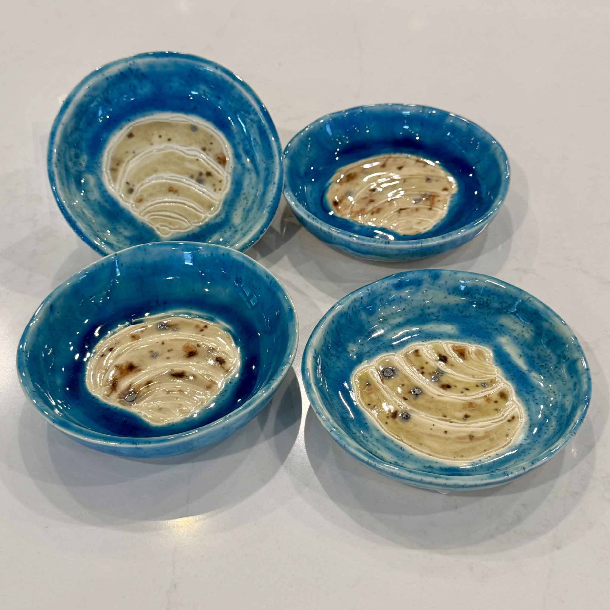 A collection of handmade Pawleys Island Shell ceramic dishes by Vida Miller displayed together, showcasing the unique variations in turquoise glaze, shell details, and handcrafted finishes. Each one-of-a-kind piece of South Carolina pottery is available at Gray Man Gallery in Pawleys Island.