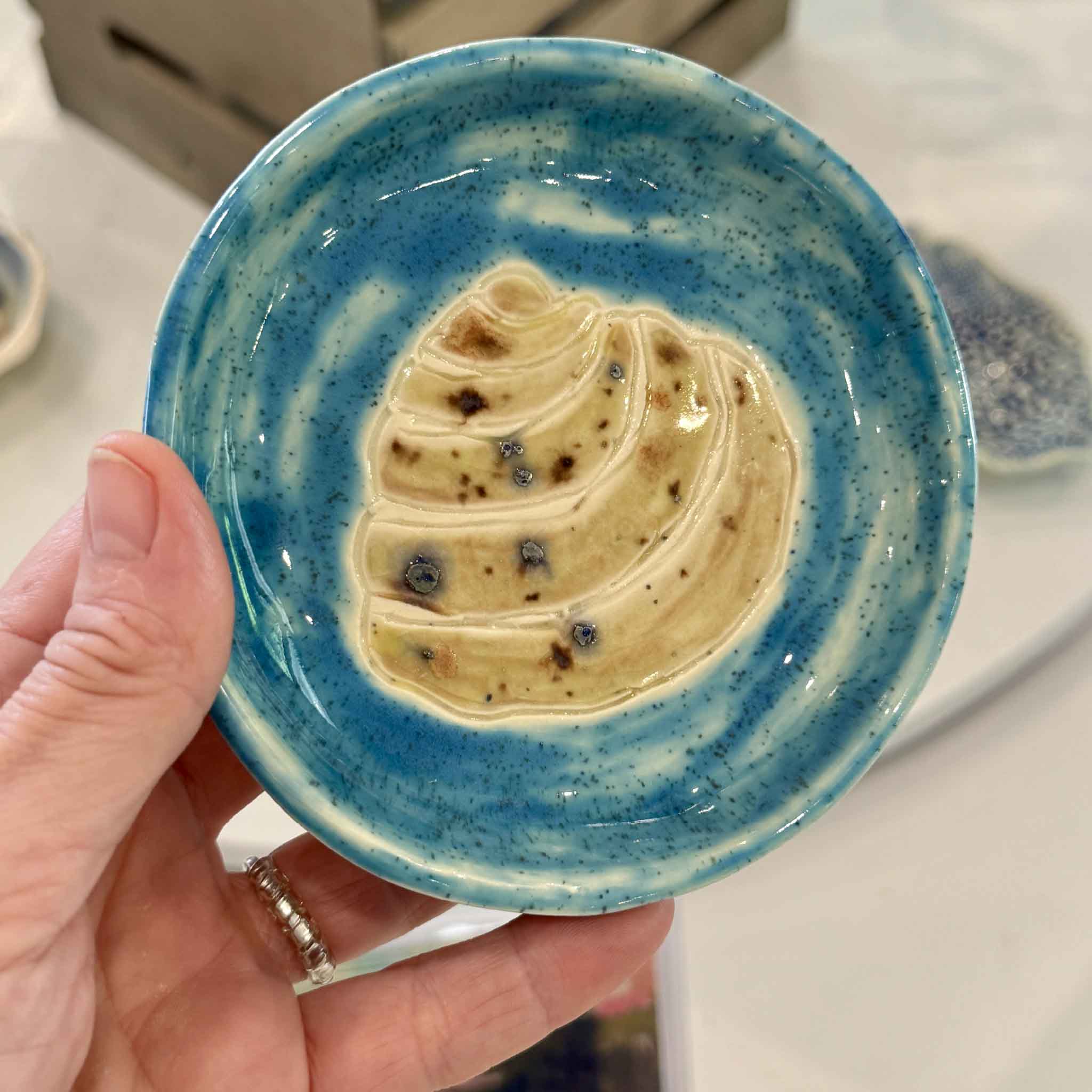 A hand holds Vida Miller's handmade Pawleys Island Shell ceramic dish finished in a vibrant turquoise glaze, showing the scale and handcrafted details of this one-of-a-kind coastal pottery piece available at Gray Man Gallery in Pawleys Island.
