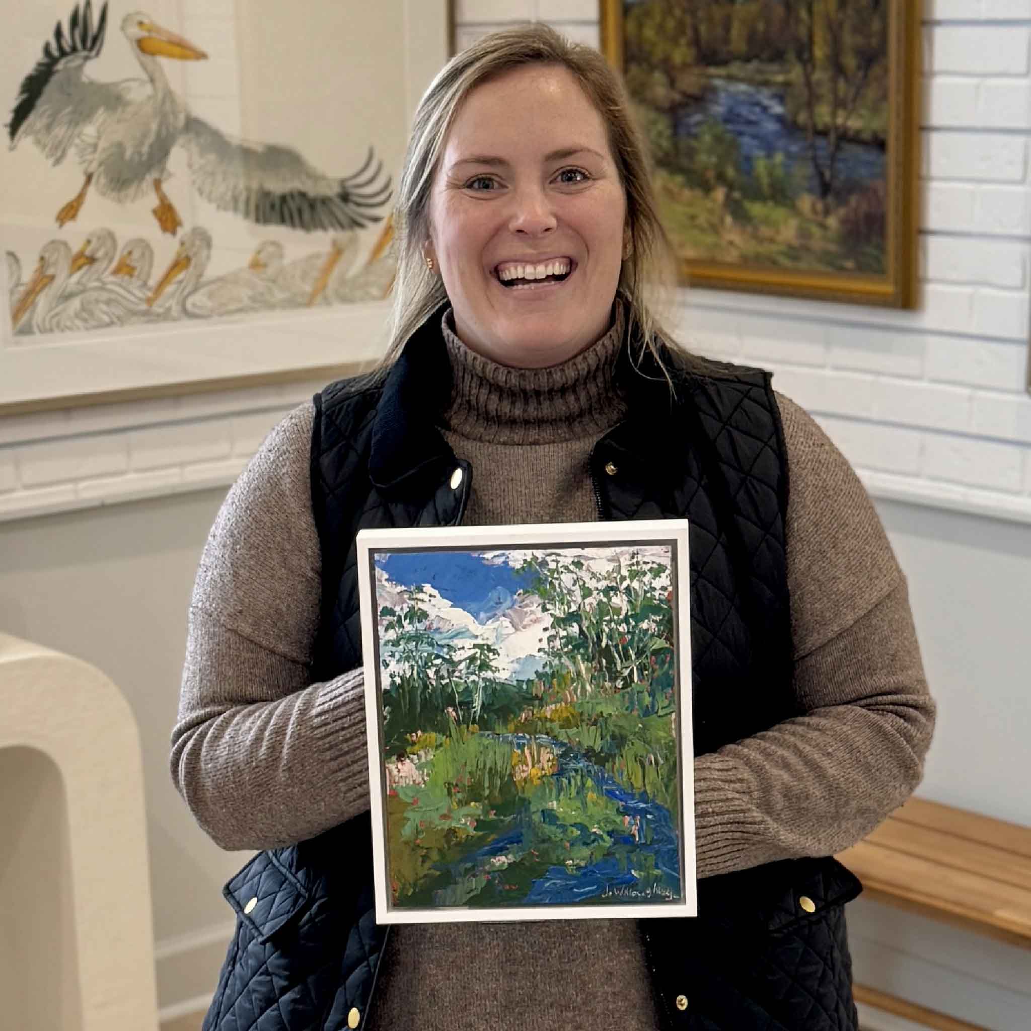 Smiling Gray Man Gallery team member holding “Wild Marsh,” an original oil painting by local artist Joanne Willoughby, featuring expressive impasto brushwork, vibrant green marsh grasses, winding blue creek, and open Lowcountry sky, photographed inside Gray Man Gallery in Pawleys Island, South Carolina, showcasing coastal fine art and custom framing craftsmanship
